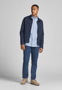 Chaqueta de botones azul marino sobre una camisa azul clara, combinada con jeans azules y zapatos beige. La chaqueta tiene un bolsillo en el pecho y un collar.