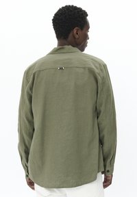Camisa de lino verde con cuello puntiagudo, puños abotonados y una etiqueta de rayas horizontales en la parte posterior. Corte entallado y mangas largas.