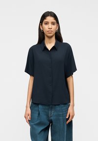 Γυναίκα που φοράει μια φαρδιά μαύρη πουκάμισα με κοντά μανίκια και πλεκτό μπλε τζιν παντελόνι, στέκεται μπροστά σε λευκό φόντο.