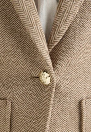 Smėlio ir kreminės spalvos striukė su įstrižai susmulkintu raštu, auksiniu mygtuku, išgraviruotu dekoratyviniu emblematu, rodoma dalis atlapų ir kišenių.