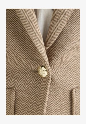 Sacou în nuanțe de bej și crem cu model herringbone, prevăzut cu un nasture auriu gravat cu un emblema decorativă, prezentând o parte din rever și buzunare.