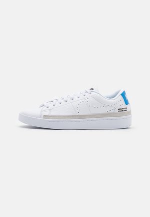 Biały niski sneaker z perforowanym logiem po boku, niebieską zakładką na pięcie, z przodu na sznurowadła oraz grubą białą podeszwą na jednolitym tle.