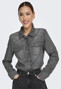 Camisa de mezclilla gris con un frente de botonadura, dos bolsillos en el pecho y mangas largas. Presenta un cuello clásico y detalles de costura.