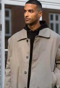 Beige yderjakke med spidse krave og sorte knapper, lagdelt over en sort hættetrøje. Glat tekstur, minimalistisk design.