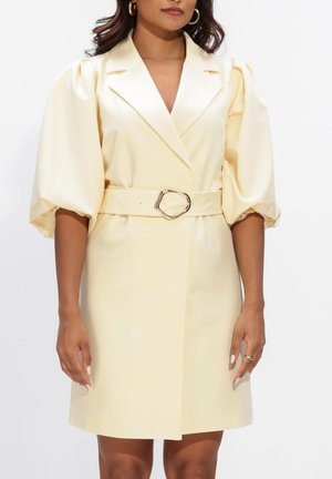 Femme portant une robe-blazer en satin crème avec des manches bouffantes et une ceinture assortie avec une boucle géométrique, debout devant un fond blanc.