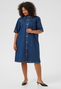 Robe chemise en denim bleu profond avec des manches courtes, fermeture par boutons et coupe droite. Équipée de coutures contrastées et d'un col classique.