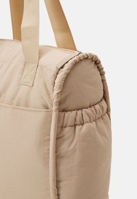 Beige vadderad duffle-väska med breda handtag och en elastisk sidoficka, visad mot en vit bakgrund.