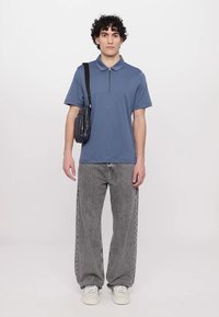 Korte ermet blå polo med halvglidelås, kombinert med lysegrå baggy jeans og hvite sneakers; modellen holder en svart skulderveske.