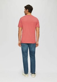 Kurzärmliges, korallenfarbenes T-Shirt aus Baumwolle, kombiniert mit dunkelblauen Jeans und weißen Turnschuhen, von hinten betrachtet.