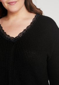 Suéter negro de punto acanalado con escote en V, adornado con un borde de encaje. La textura es suave y ligeramente transparente, resaltando el detalle del escote.