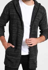 Cardigan con cappuccio in un tessuto lavorato in nero e grigio. Presenta una cerniera, maniche lunghe e due tasche frontali. Indossato sopra una t-shirt bianca.