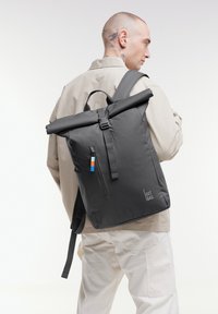 Sac à dos à rabat gris avec des bretelles noires, poche avant zippée mettant en valeur des accents bleus et orange, fabriqué à partir de matériaux durables. Logo de la marque affiché.