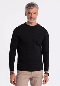 Musta pitkähihaisa t-paita slim fit -mallissa, valmistettu puuvillasta, jossa on pyöreä pääntie ja sileä pinta. Yhdistetty vaaleanbeigeihin housuihin.