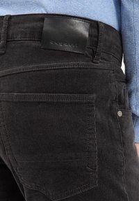 Black corduroy pants with ribbed texture, featuring a leather patch at the waist and standard pockets. Close-up on back side.