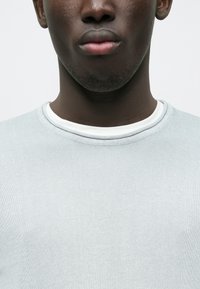 Sweatshirt cinzento claro com tecido texturizado e de malha. Apresenta uma gola redonda sobreposta com uma gola interior branca.