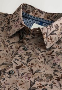 Camicia marrone con motivo floreale, caratterizzata da un tessuto testurizzato, apertura con bottoni e un colletto interno con motivo a contrasto. I dettagli includono bottoni piccoli.