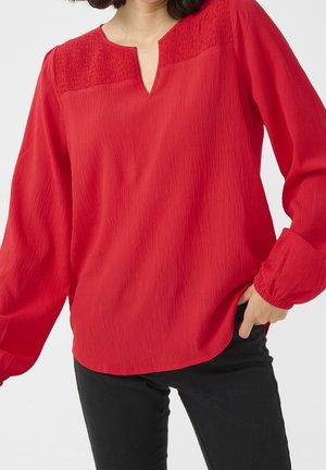 Femme portant un chemisier rouge vif à manches longues avec un empiècement texturé et un pantalon noir, debout avec une main dans la poche.