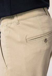 Beige chinos med en slät textur, rak skärning och elastisk midja. Har en bakficka med en knapp och en subtil sömdetalj.