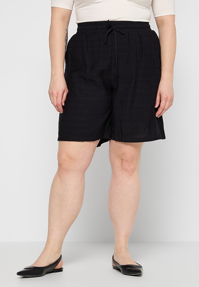 Frau in schwarzen knielangen Zugbandshorts und schwarzen flachen Slingback-Schuhen, die vor einem schlichten Hintergrund steht.