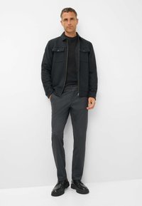 Donkergrijze ritsjack met twee borstzakken over een zwarte trui, gecombineerd met lichtgrijze broek en zwarte schoenen, tegen een witte achtergrond.