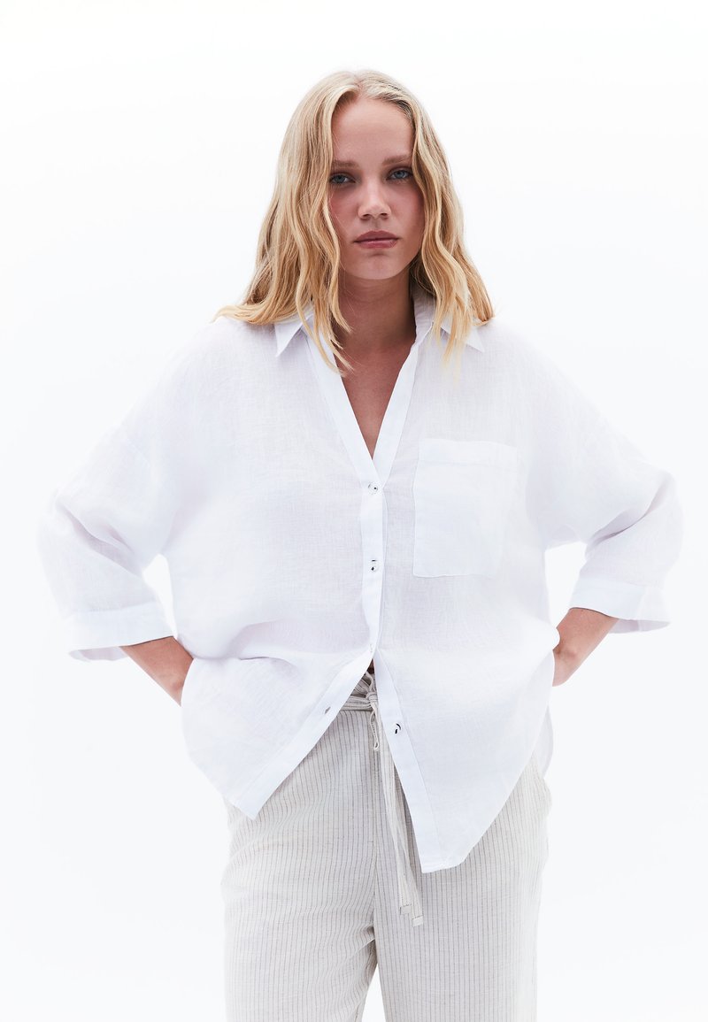 White linen button-up shirt with a relaxed fit, featuring a chest pocket and rolled sleeves, paired with light ribbed pants.
