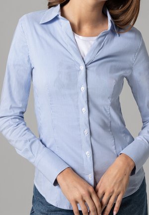 Mujer con una camisa entallada de botones azul claro a rayas finas sobre una camiseta blanca, con las manos entrelazadas al frente, sobre un fondo gris claro liso.