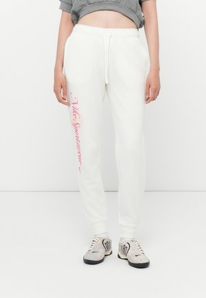 Persoon draagt witte sweatpants met trekkoord en roze "Nike Sportswear"-tekst op linkerbeen en lichtgrijze sneakers met zwarte zolen.