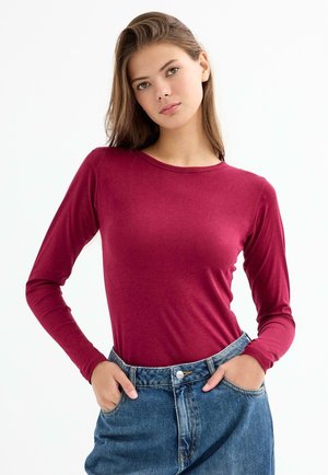 Camisa de manga larga color burdeos, confeccionada en tela suave, con cuello redondo y mangas ajustadas, combinada con shorts de mezclilla de talle alto.