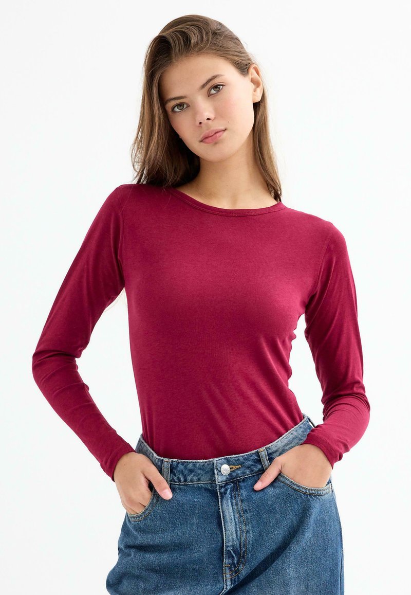 Camisa de manga larga color burdeos, confeccionada en tela suave, con cuello redondo y mangas ajustadas, combinada con shorts de mezclilla de talle alto.