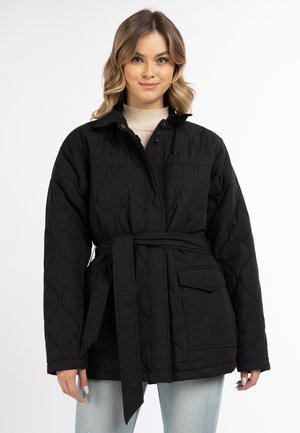 Frau trägt eine schwarze gesteppte Jacke mit Gürtel, Knopfleiste vorne, großer Tasche und hellblauen Jeans vor einem schlichten weißen Hintergrund.
