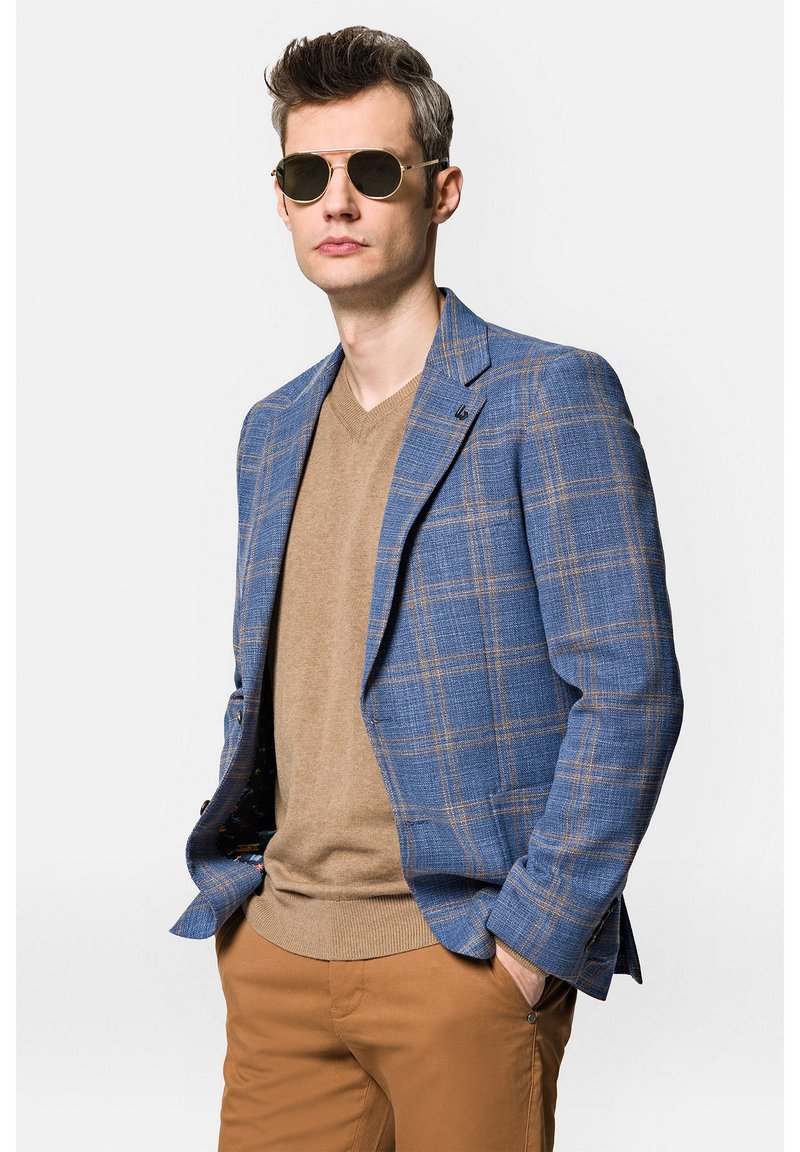 Niebieska marynarka w kratę z brązowymi akcentami, kremowy sweter z dekoltem w serek i jasnobrązowe spodnie. Model w okularach przeciwsłonecznych, ręce w kieszeniach.