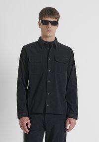 Camisa negra de botón con dos bolsillos en el pecho, hecha de tela texturizada. Cuenta con un cuello y puños, combinada con pantalones a juego y gafas de sol.