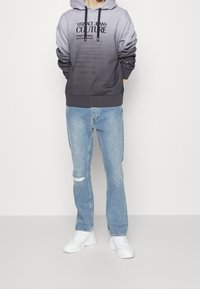 Mikina s kapucí v šedém přechodovém odstínu, zdobená textovými motivy. Kombinována s světle modrými džínami, jedny s trhlinou na koleni, a bílými teniskami.