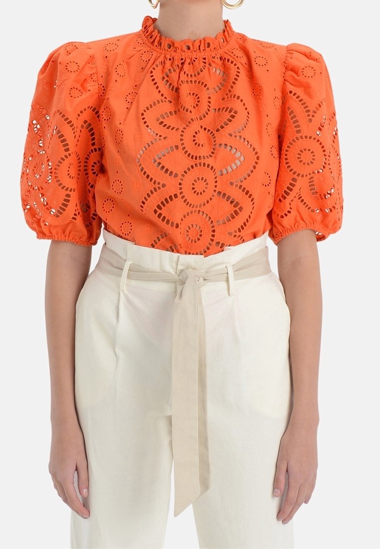 Oranje blouse met oogjes en pofmouwen, verzamelde halslijn, bloemen uitsneden patroon. Gecombineerd met hooggesneden crèmekleurige broeken en een stoffen riem.