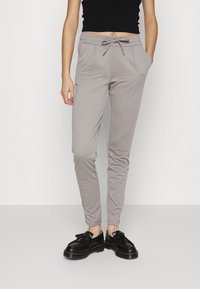 Calças de jogging cinza claro com cintura elástica e cordão. Possui bolsos laterais e um ajuste slim, combinadas com sapatos pretos.