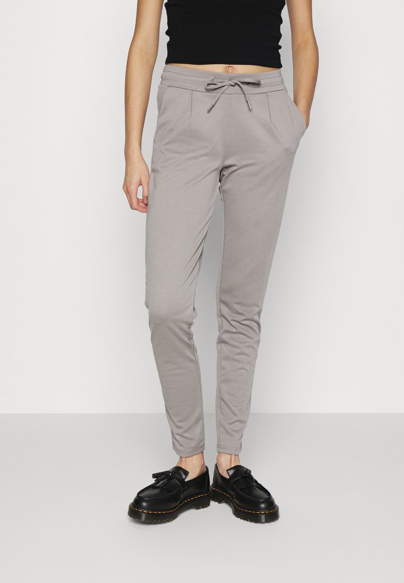 Calças de jogging cinza claro com cintura elástica e cordão. Possui bolsos laterais e um ajuste slim, combinadas com sapatos pretos.