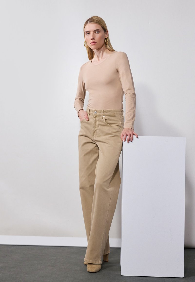 Beige long-sleeve top paired with high-waisted, light khaki wide-leg pants. Model stands beside a white, square pedestal with a matte finish.