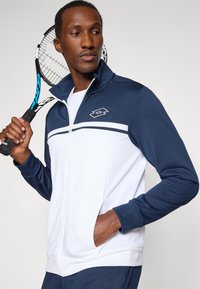 Blaue und weiße Zip-Up-Trainingsjacke mit hohem Kragen, mit einem Logo auf der Brust und gestreiftem Design. Das Model hält einen Tennisschläger.