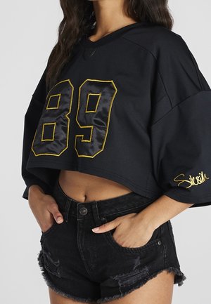 Frau trägt schwarzen, kurz geschnittenen, übergroßen Pullover mit der gelb umrandeten Zahl 89 und schwarzen, abgenutzten Jeansshorts, Hände in den Taschen.