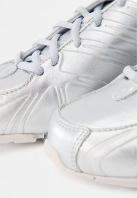 Ασημένια αθλητικά παπούτσια με λεία δερμάτινη υφή, σχεδιασμένα με κορδόνια και διακριτές καμπύλες στην πάνω επιφάνεια. Λευκή καουτσούκ σόλα.