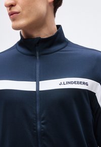 Άνδρας που φοράει μπουφάν με φερμουάρ σε ναυτικό μπλε χρώμα με λευκή οριζόντια ρίγα και κείμενο "J.LINDEBERG" στο στήθος.