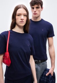 Δύο νέοι ενήλικες φορούν μπλε μπλουζάκια ναυτικού χρώματος· η γυναίκα έχει μία κόκκινη τσάντα ώμου, ο άνδρας κρατάει ένα καρό μπλε σακάκι, στεκόμενοι μπροστά σε απλό φόντο.