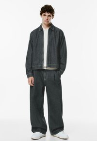 Dark denim jacket with a zip closure, paired with matching loose-fit pants. Both have a smooth texture, worn over a light-colored top.