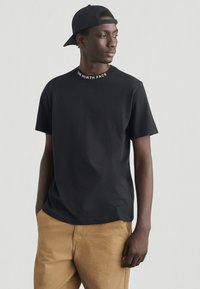 Μαύρο t-shirt με κοντά μανίκια και λευκό λογότυπο "THE NORTH FACE" στον γιακά, συνδυασμένο με καμηλό παντελόνι. Καπέλο φορεμένο προς τα πίσω. Λείο ύφασμα.