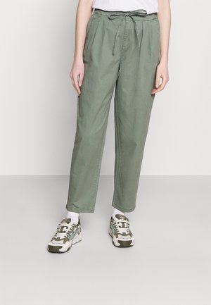 Personne portant un pantalon taille haute vert clair avec une taille à cordon, des chaussettes blanches et des baskets blanches et vertes épaisses, debout sur un sol gris.