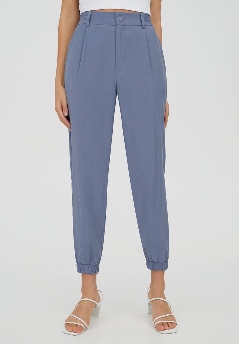 Femme portant un pantalon bleu taille haute, fuselé, avec des poignets élastiques et des sandales à talons blanches à bout ouvert, debout devant un fond uni.