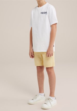 Joven con una camiseta blanca con la palabra "Paradise", pantalones cortos beige, calcetines blancos y zapatillas blancas, posando contra un fondo liso.