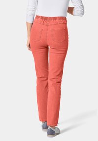 Koraalkleurige jeans met elastische taille, rechte snit, vijf zakken en een soepele denim textuur. Gedragen met grijze veterschoenen.