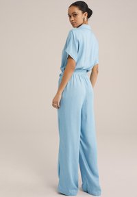 Lyseblå jumpsuit med korte ærmer, brede ben og en tætsiddende talje. Blødt stof med en glat tekstur; minimal detaljer.