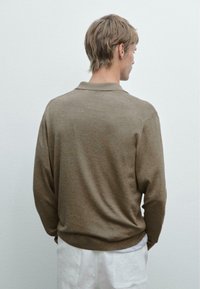 Hnědá polo košile s dlouhým rukávem a límečkem, která má volný střih a texturovaný pletený vzor. Nosená s volnými, světlými šortkami.